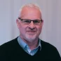 Smiling man with glasses, in a collared shirt with a black sweater over it.