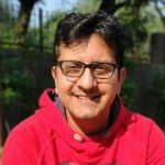 Nitin in a read sweater and glasses against a nature backdrop