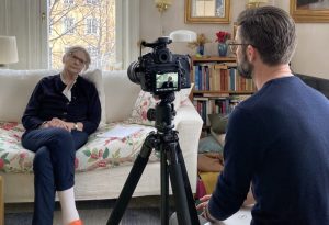 Malin Falkenmark sitting on a floral printed white couch, being shot through a lens on a tripod behind which Andreas Karlsson is sitting with his back in the photo.