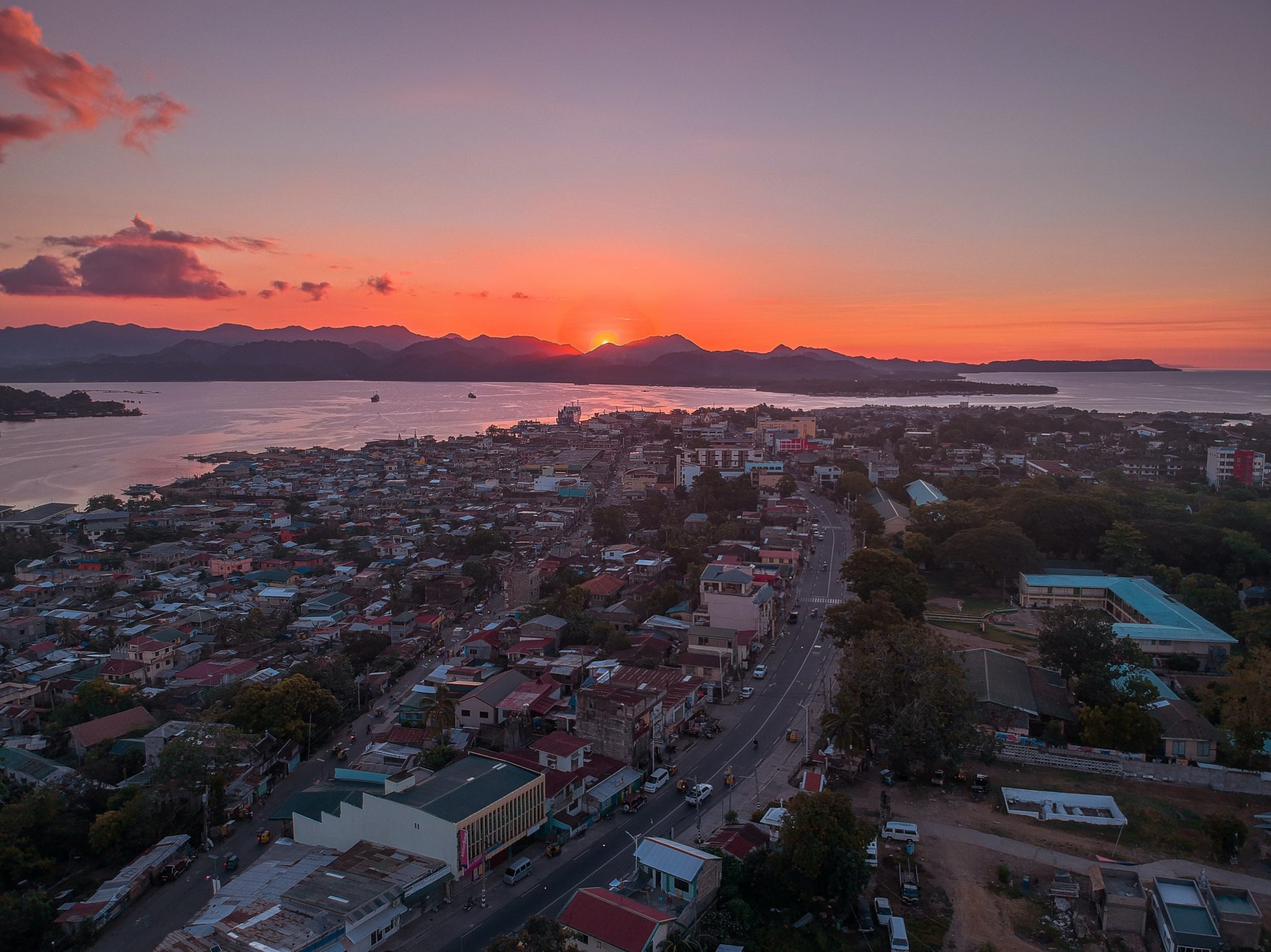 Aerial view of a city with small houses, by a river. The sun is rising in the background