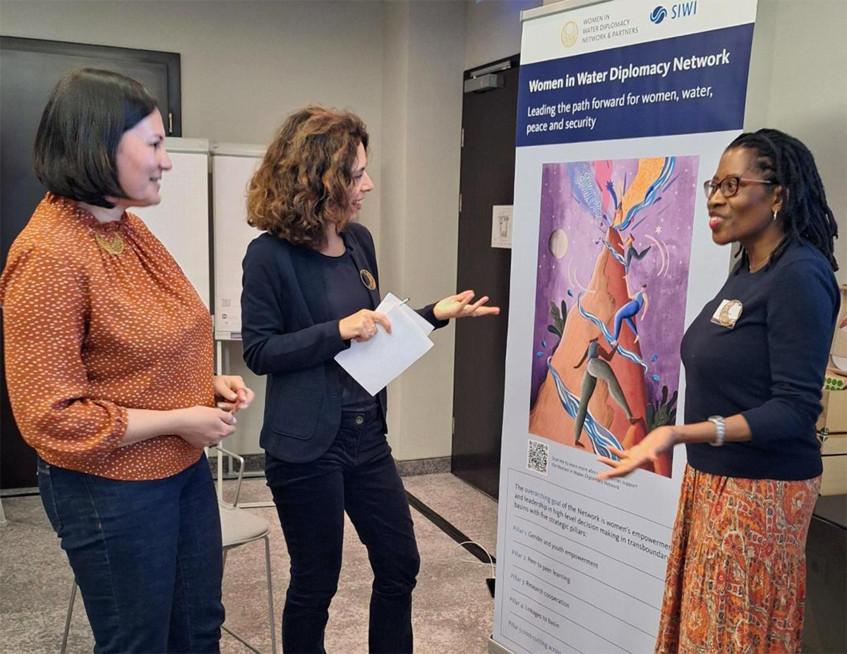 3 women - members of the Women in Water Diplomacy Network - standing in front of the banner showing the illustration "you raise me up".