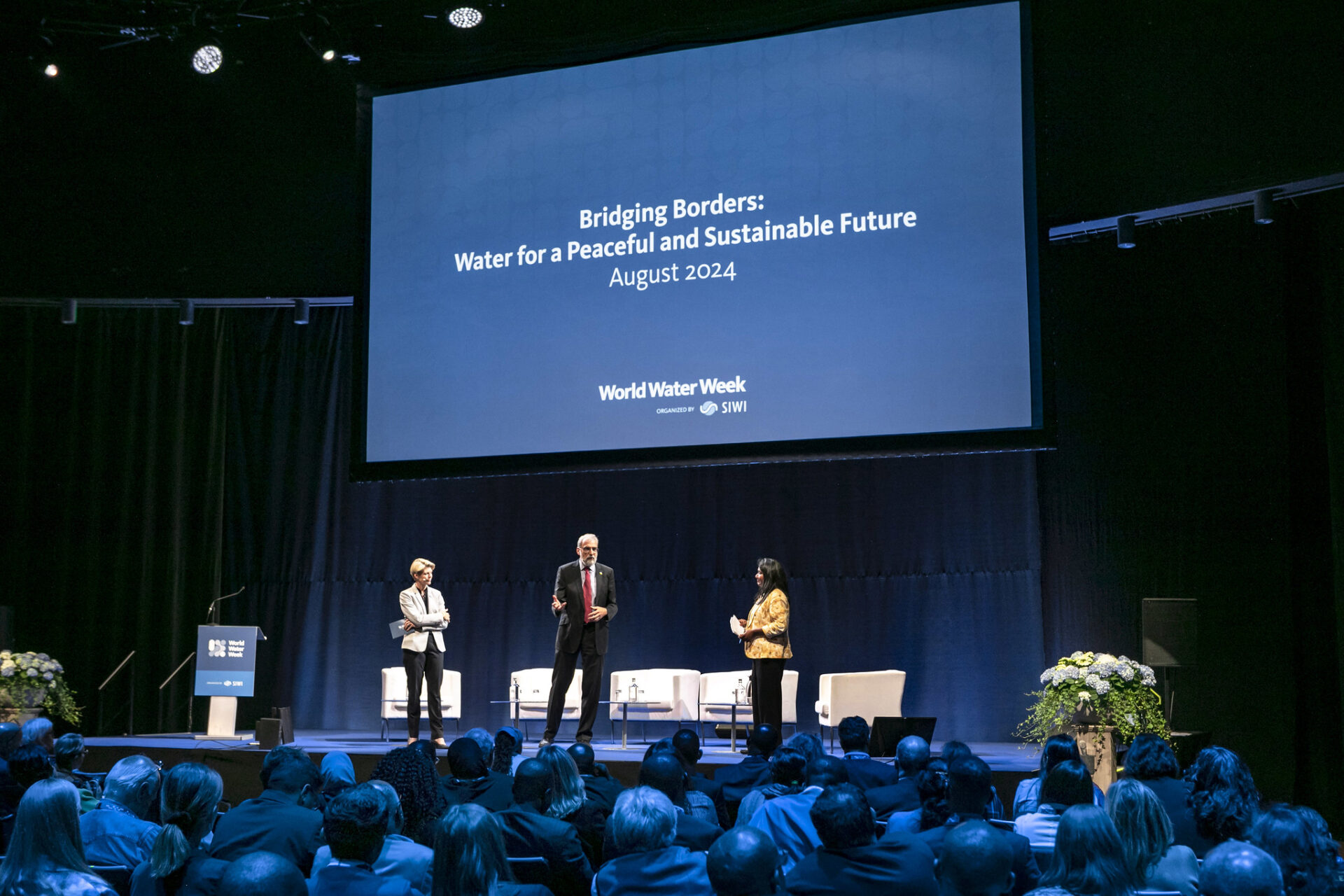 Jone Lane in the middle with 2 people on either side of the World Water Week stage with audience lit up by blue lights watching over. The blue screen on stage reads Bridging Borders: Water for a Peaceful and Sustainable Future.